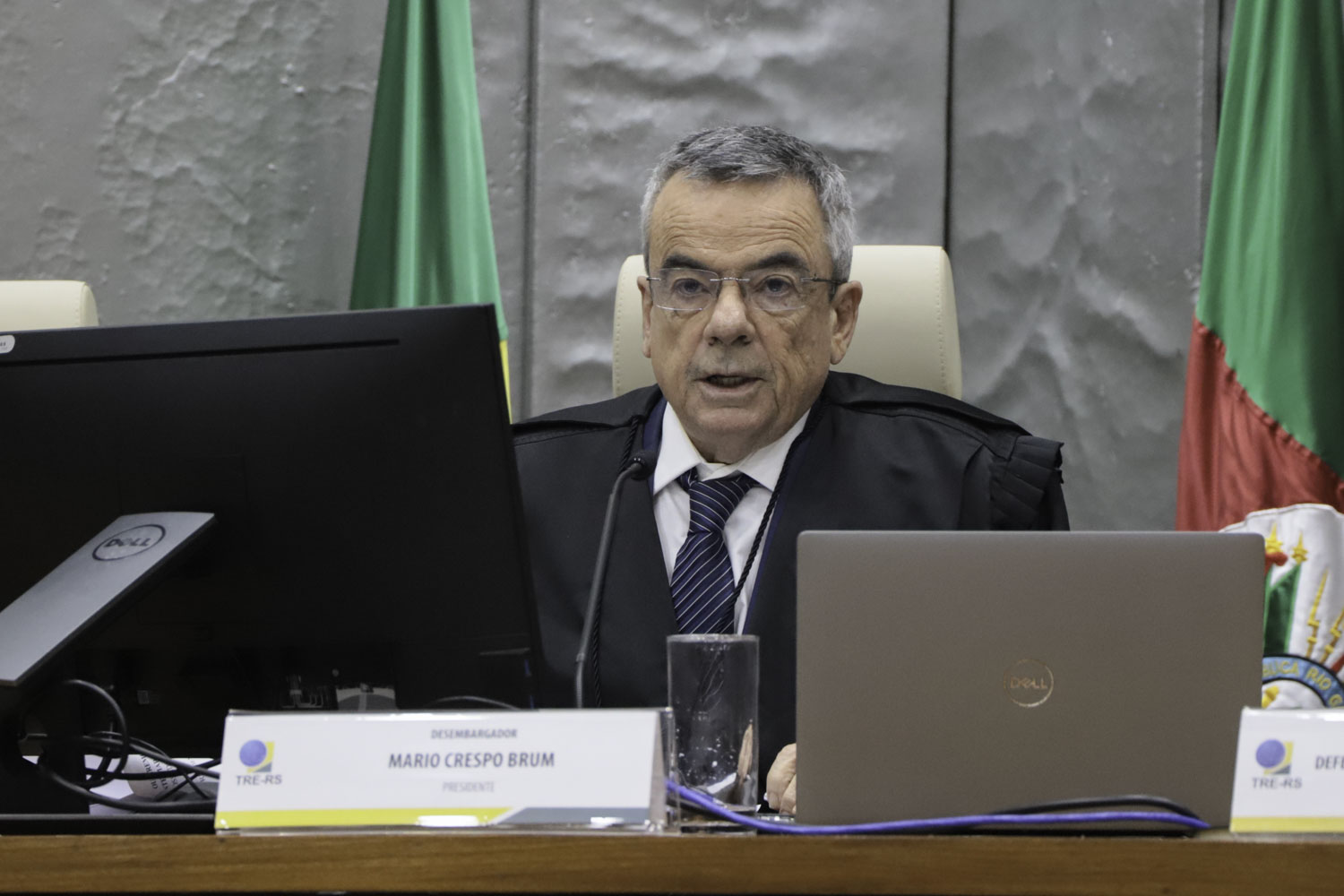 Mario é branco e grisalho. Usa óculos de aro fino e fala o microfone. Veste toga, camisa branca e gravata azul e preta. 