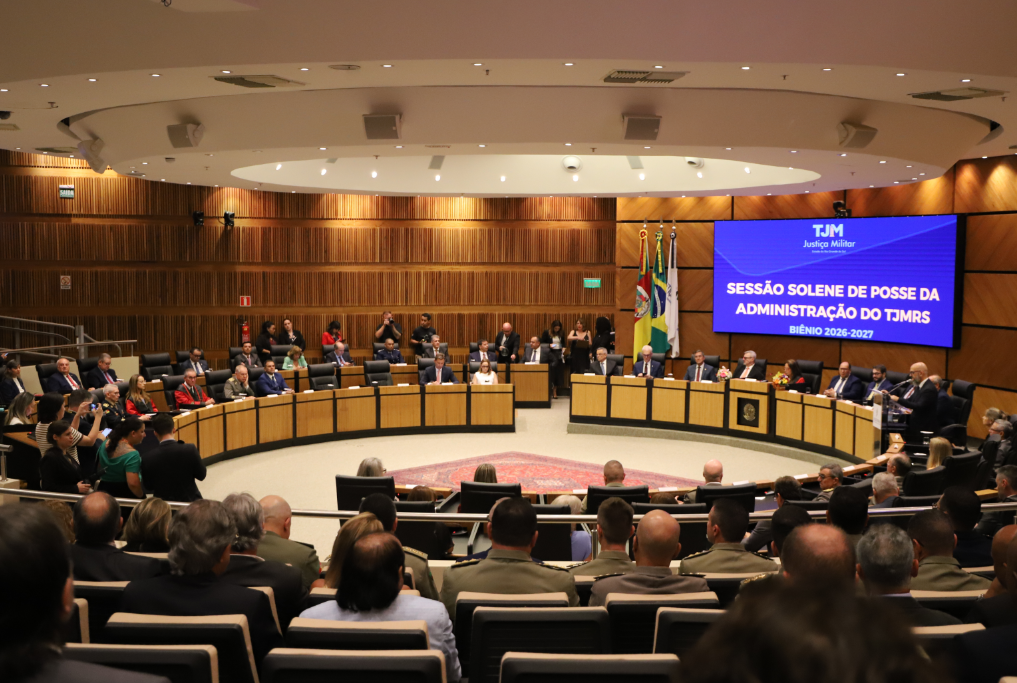 Vista panorâmica do auditório durante a Sessão Solene de Posse do TJMRS. Autoridades ocupam uma longa bancada em "U", posicionada à frente de um grande telão azul com o título do evento e bandeiras oficiais. O teto possui um design circular com iluminação embutida. Em primeiro plano, a plateia é vista de costas, composta por civis e diversos militares fardados.