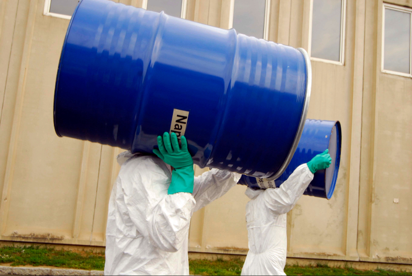 Dois trabalhadores usando Equipamentos de Proteção Individual (EPIs) carregam uma bombona com produtos químicos.