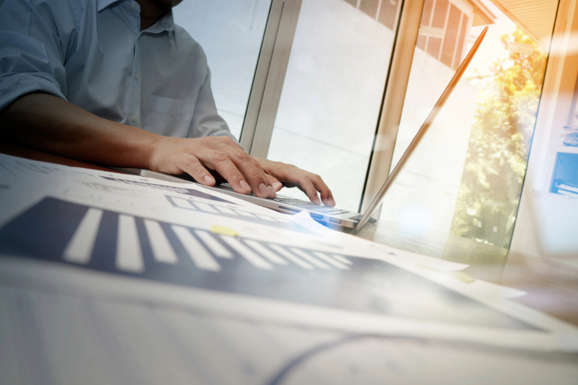 Homem digita em notebook. Sobre a mesa, há documentos com planilhar financeiras. Ele veste camisa azul clara e não há imagem do rosto.