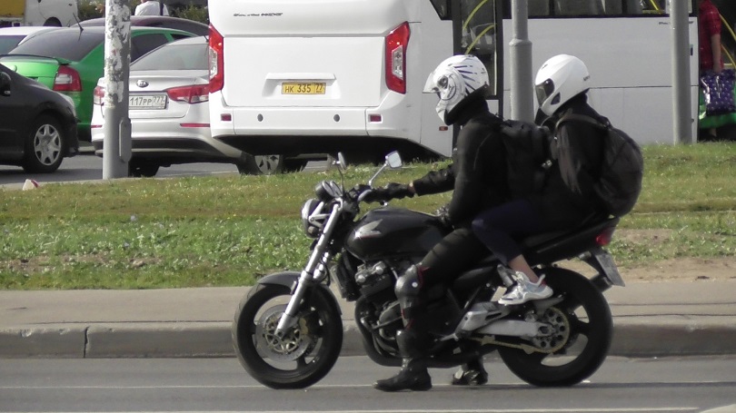 Foto ilustrativa que retrata uma motocicleta preta sendo conduzida em uma via urbana, transportando duas pessoas.