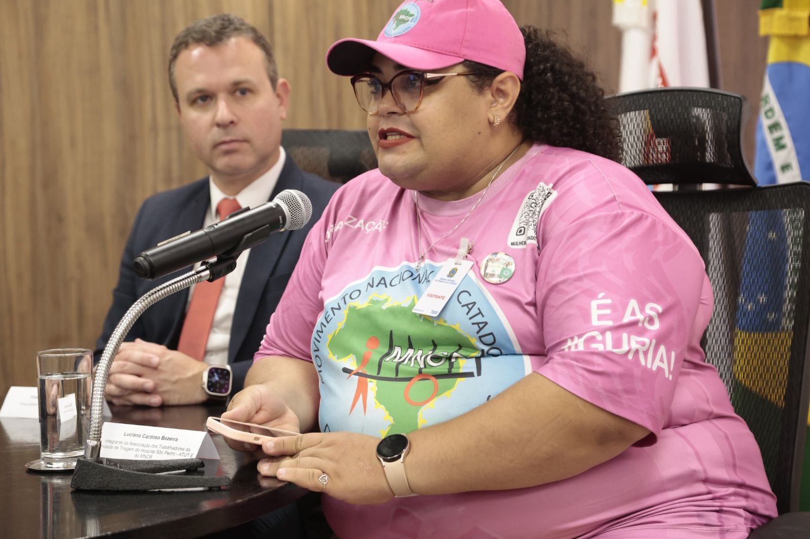 Luciana é negra de cabelos cacheados escuros. Usa bone e camiseta rosa, com o logo do movimento naicional dos catadores e catadoras. 