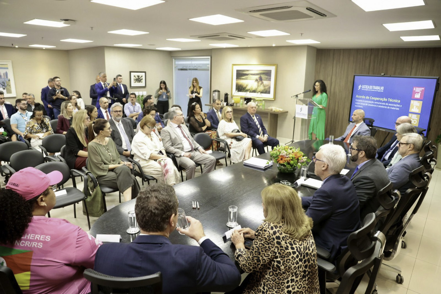foto geral do evento, com o público à esquerda, a mestre de cerimônias ao centro e a mesa oficial à direita. 