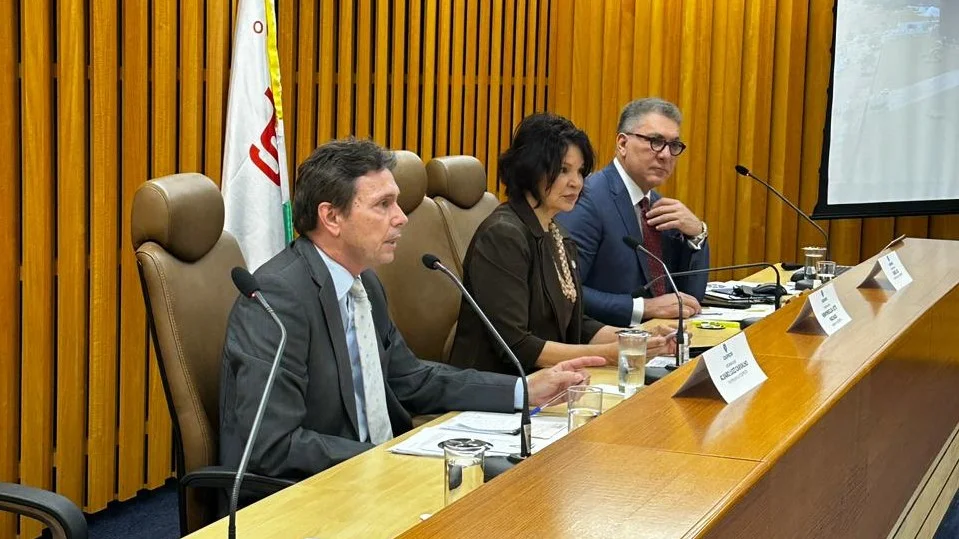 mesa oficial com três pessoas de perfil. Um homem em primeiro plano fala ao microfone. 