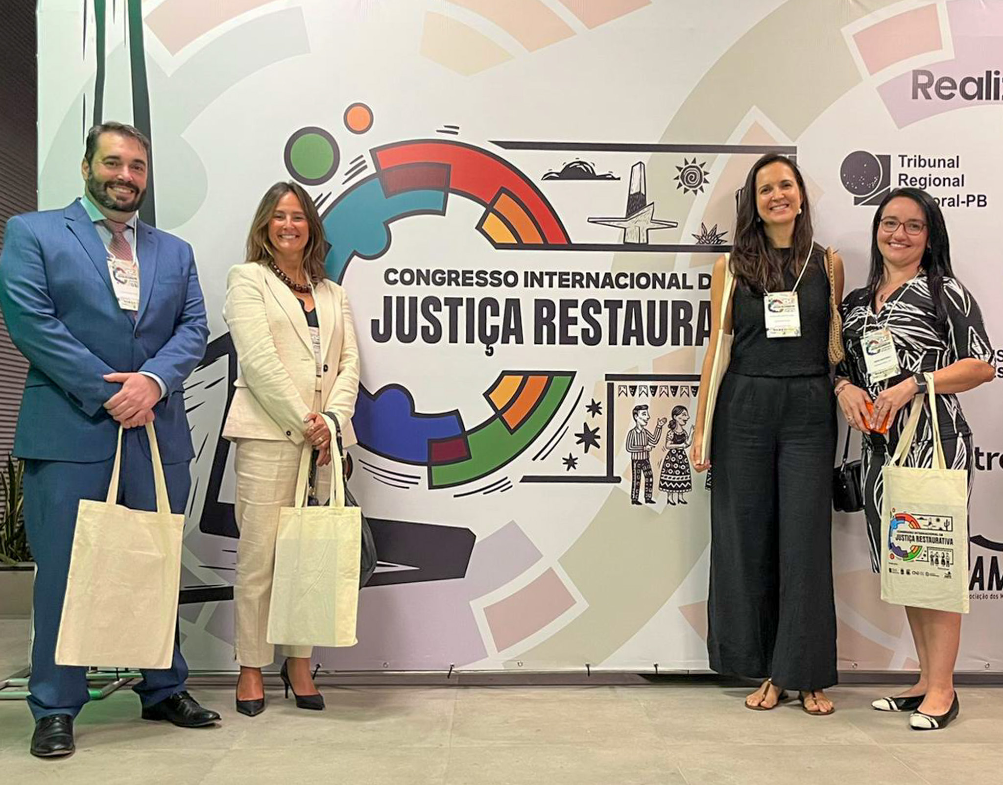 Quatro pessoas posam sorrindo em frente ao painel do Congresso Internacional de Justiça Restaurativa, segurando bolsas do evento, em um espaço interno de convenções.