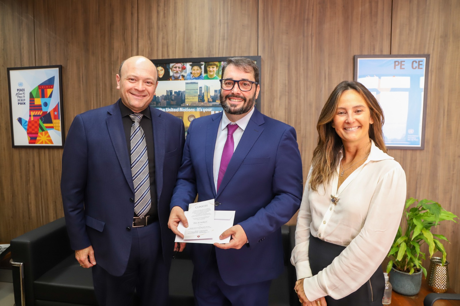 A foto mostra três pessoas lado a lado posando para foto, sendo a do meio com ele em mãos.