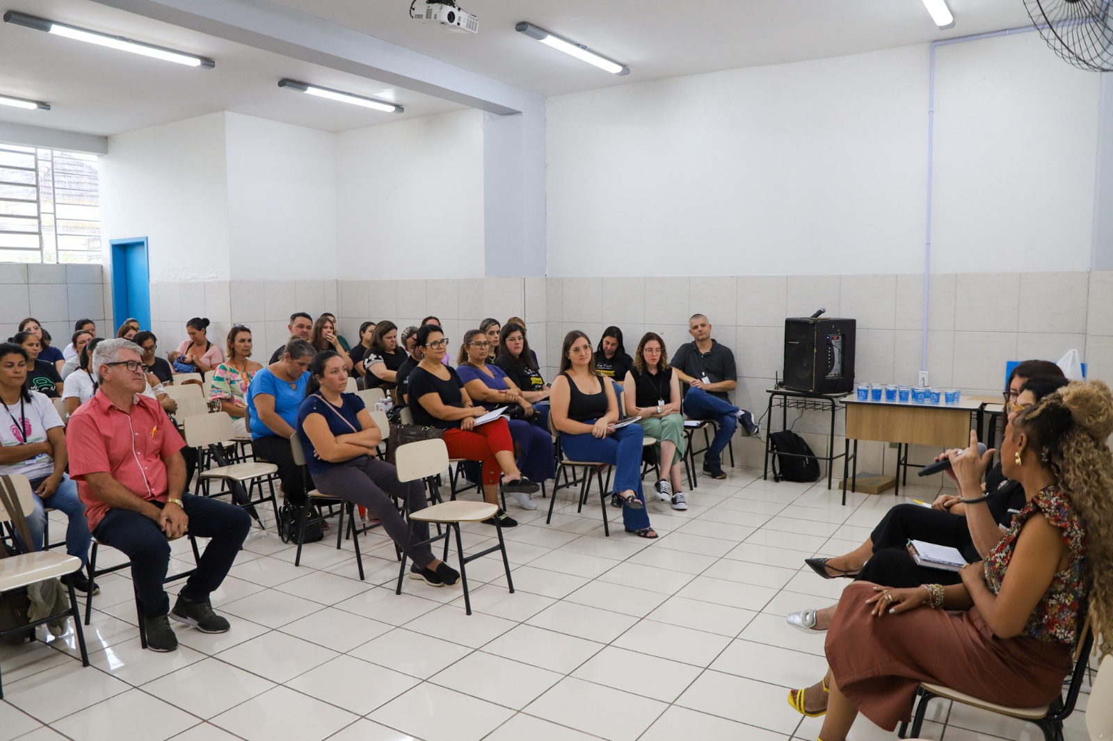 A foto mostra a desa. Brigida falando de lado e público assistindo.
