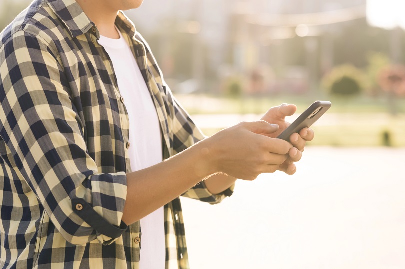 Foto ilustrativa. Homem em pé ao ar livre usando celular, vestindo camisa xadrez sobre camiseta branca, com luz do sol e fundo desfocado que parece um parque ou praça.