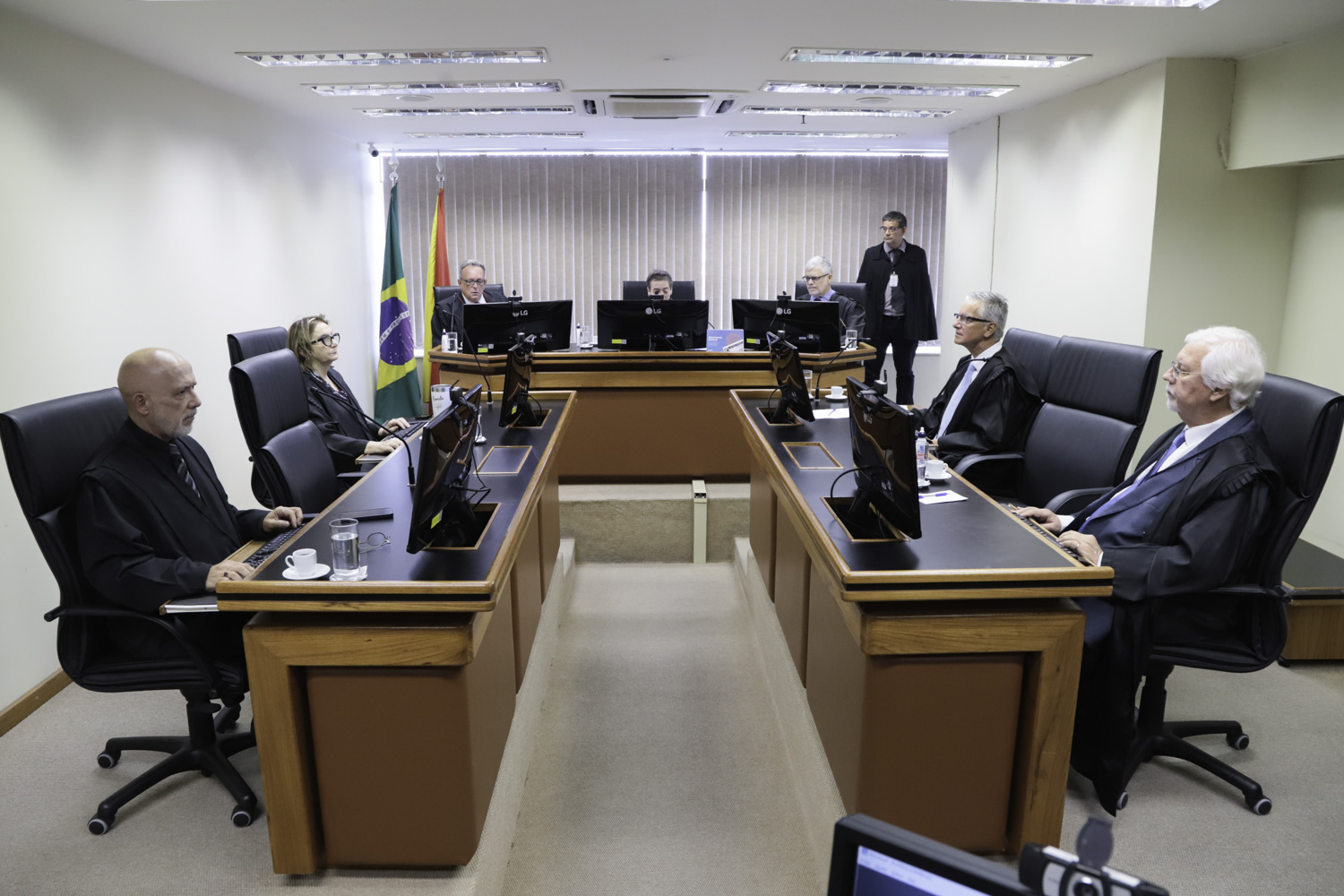 oito pessoas aparecem na imagem, durante a sessão de julgamento. Os magistrados estão em mesas nas laterais e ao fundo, em formato de U.