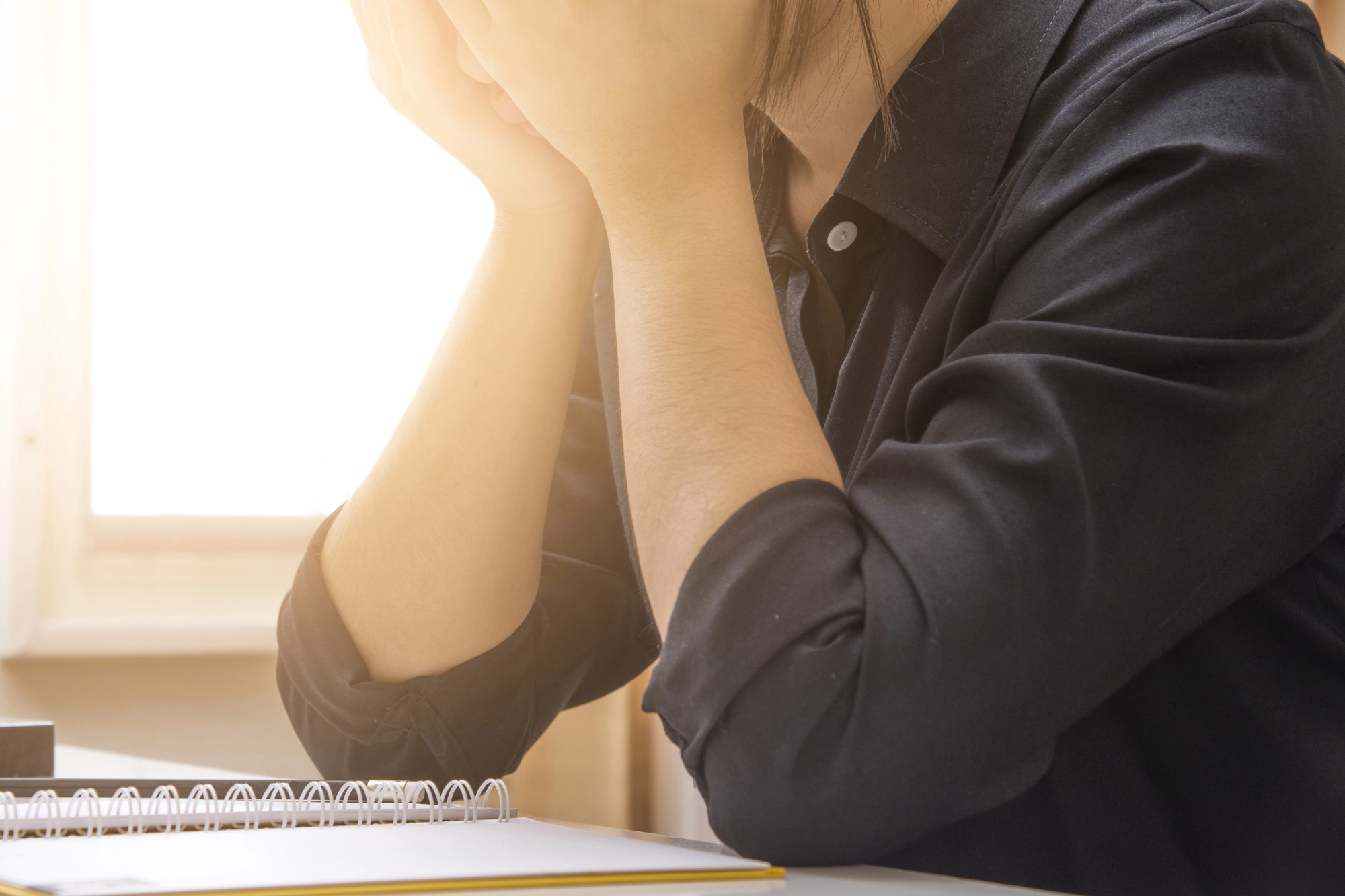 Mulher com as mãos no rosto em frente a uma mesa de trabalho com um caderno aberto.