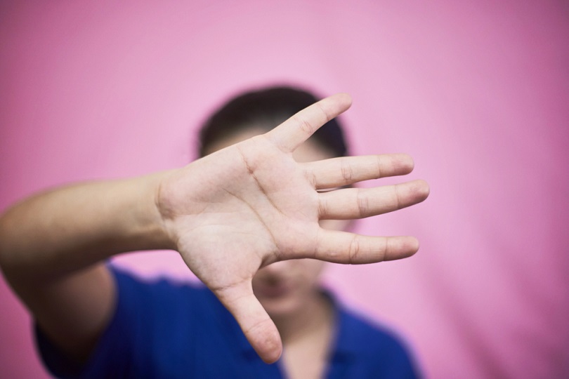 Mulher diante de um fundo rosa levanta a mão aberta em direção à câmera, cobrindo o rosto em sinal de proteção, recusa ou pedido de ajuda contra a violência.