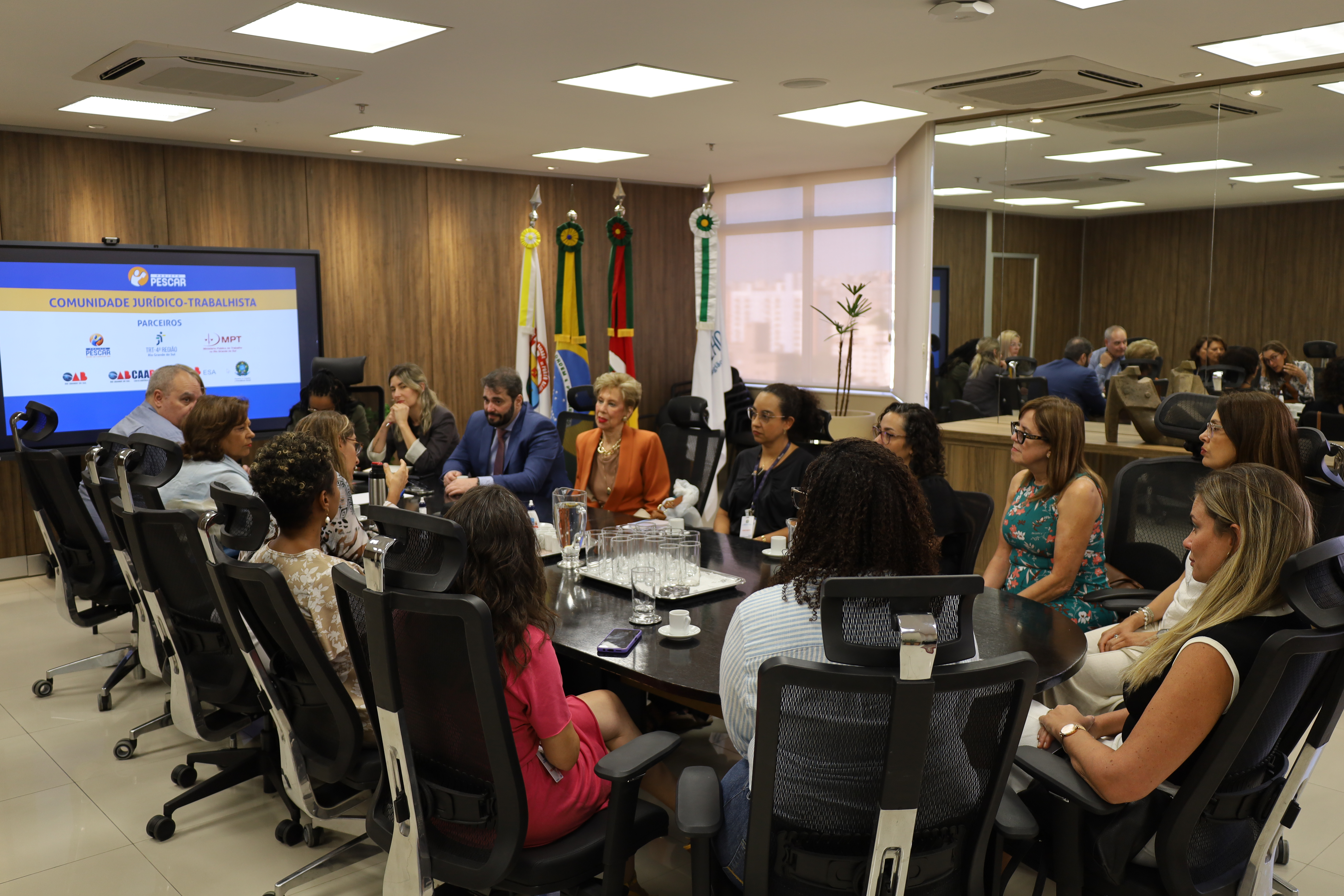 Em outro ângulo da mesma sala, os participantes aparecem distribuídos ao redor da mesa oval, em cadeiras pretas de escritório. A tela ao fundo projeta a logo do Projeto Pescar e dos parceiros. Algumas pessoas falam enquanto outras escutam com atenção, com anotações e celulares sobre a mesa.