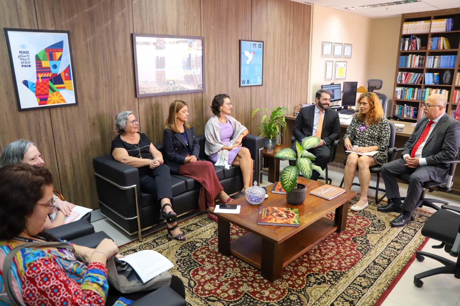 A foto mostra pessoas sentadas em poltronas e cadeiras numa reunião, numa sala fechada bem iluminada.