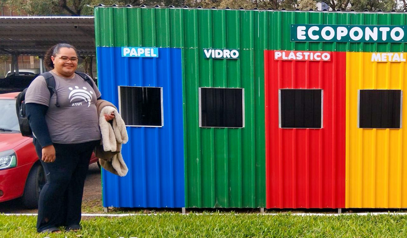 Luciana posa sorrindo ao lado de um ecoponto colorido feito de contêiner metálico. Cada abertura está identificada por cor e material: azul para papel, verde para vidro, vermelho para plástico e amarelo para metal. 