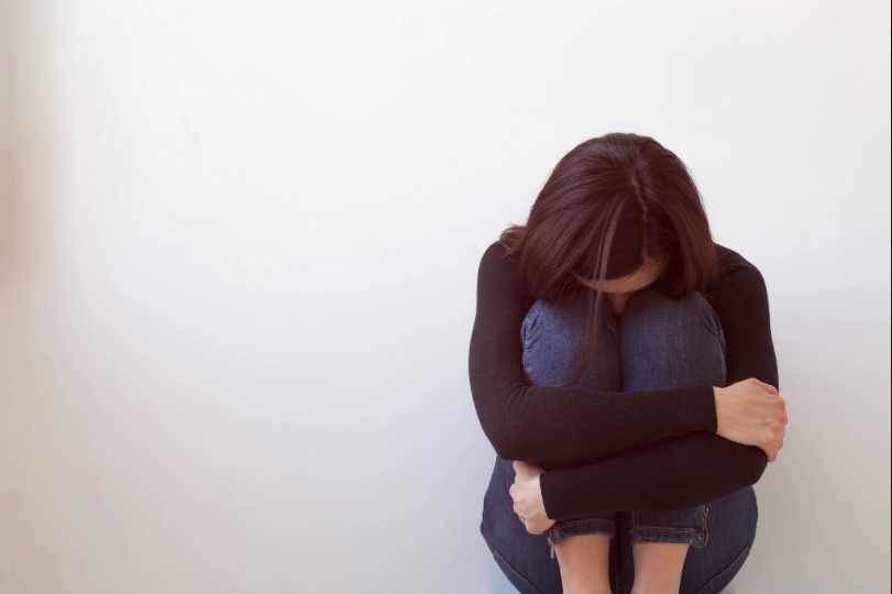 Mulher abraça os joelhos e apoia a cabeça sobre eles. Ela tem a pele branca e cabelos pretos e lisos. Usa blusa preta e calça jeans escura. O fundo da imagem é branco.