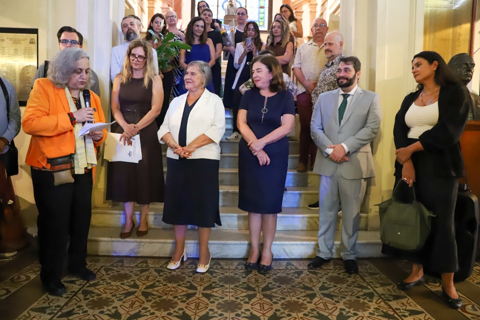 A foto mostra evento em homenagem às mulheres na Ufrgs. Cerimônia com uma pessoa falando ao microfone e outras ao lado.