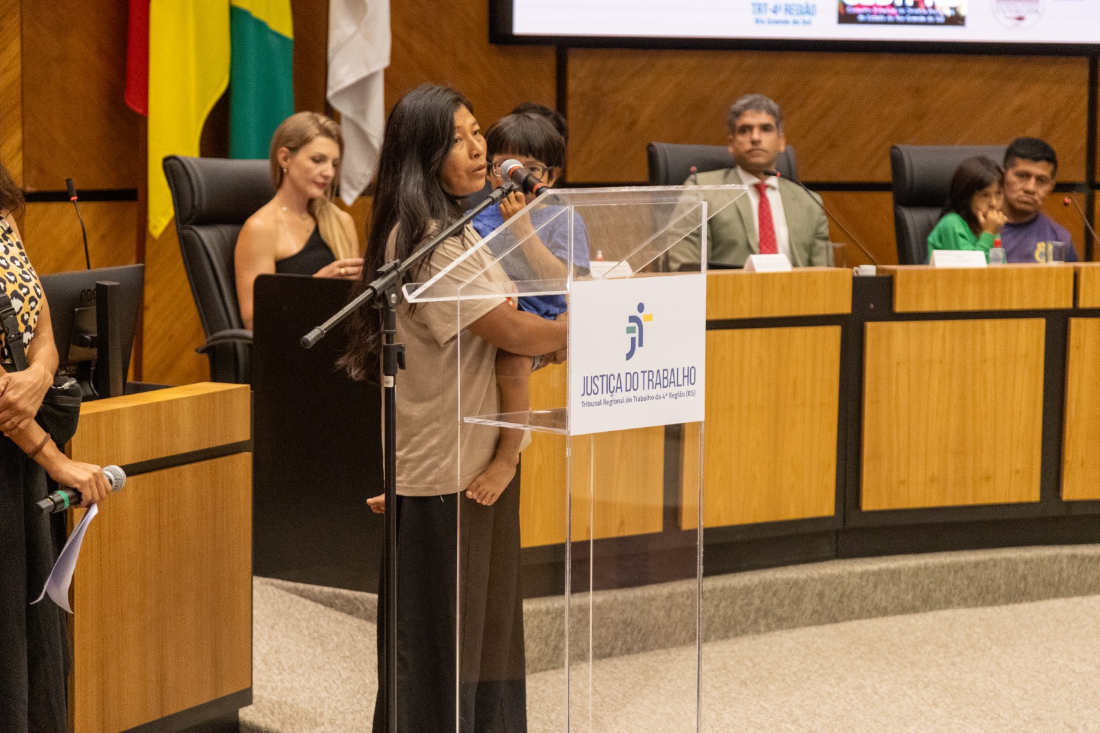 Patrícia fala ao microfone com criança no colo. Ela é indígena, tem cabelo preto liso e comprido.  