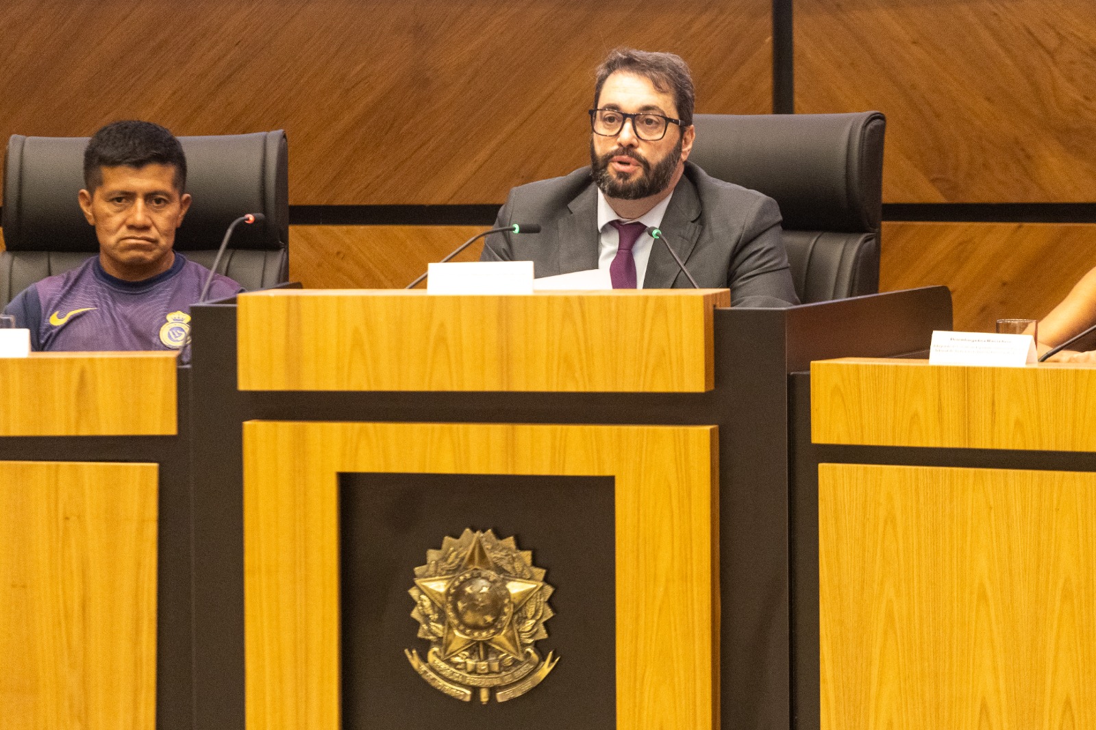 Aldo é indígena, de cabelo preto escuro, e Alexandre é branco, de cabelo e barba escura. Os dois estão na mesa oficial do evento. 