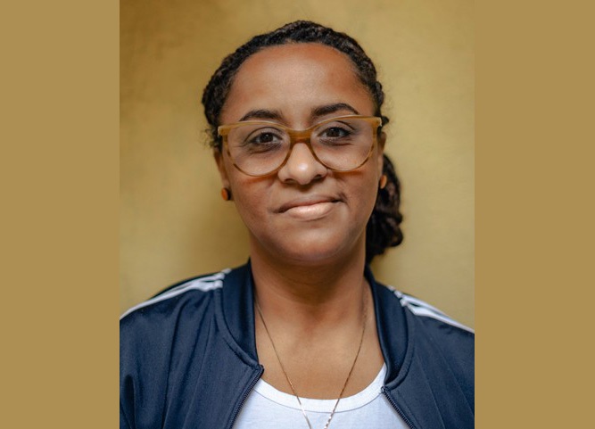 Lília é negra. usa cabelo cacheado escuro preso e jaqueta azul com listras brancas nos ombros. usa óculos de aro grande e está sorrindo.