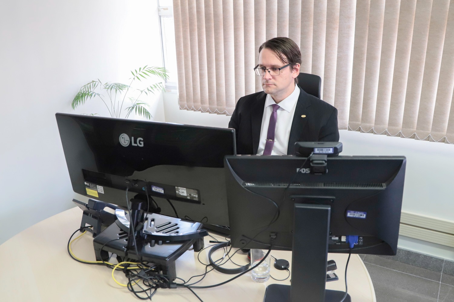Fabrício é branco, de cabelo liso e castanho. está de terno e gravata, em sua mesa de tabalho. Olha para duas telas de computador. 