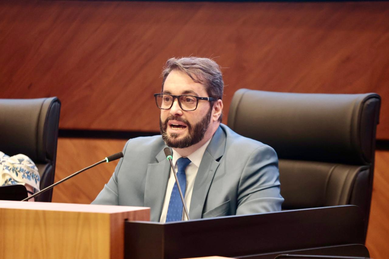Alexandre é branco, de barba e cabelos pretos. Usa óculos de aro preto e veste terno azul, com camisa e gravata azul. fala ao microfone 