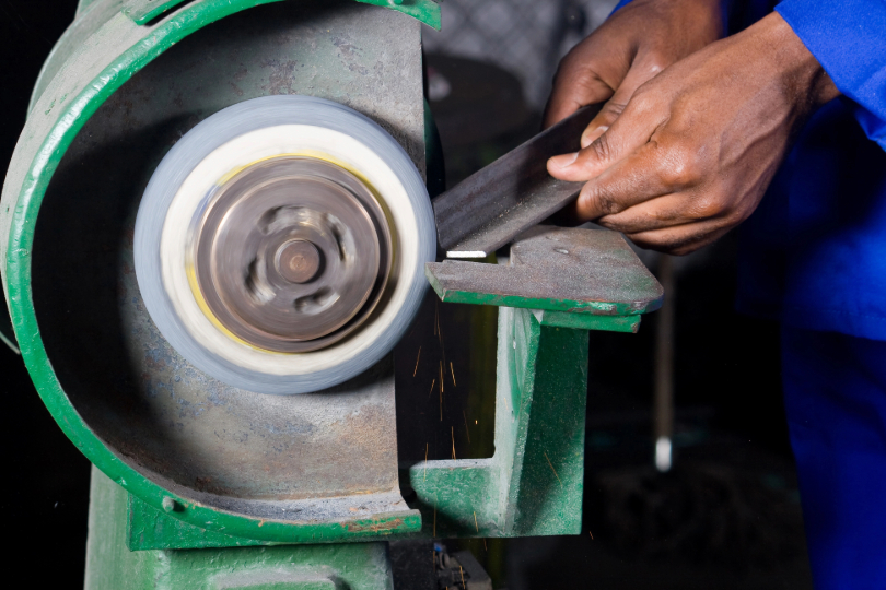 Mão de homem negro aproxima objeto de metal de máquina industrial. A máquina é verde e ele usa uniforme azul.