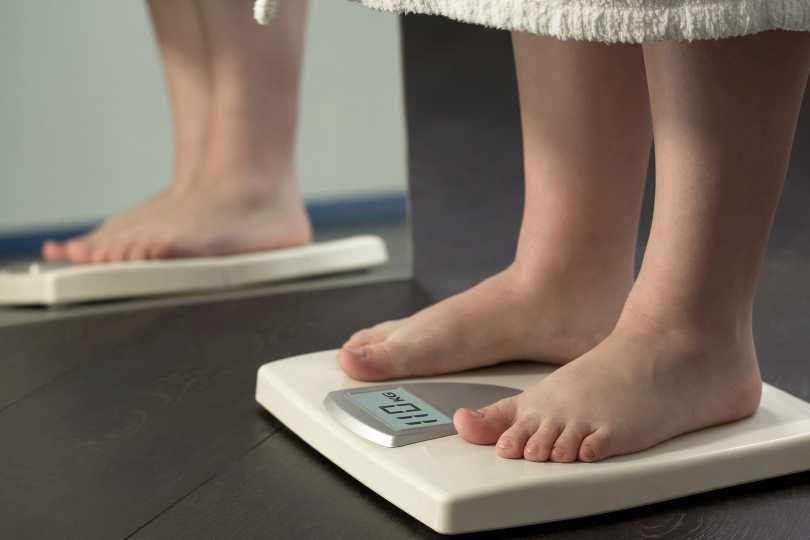 imagem de uma mulher sobre balança refletida em espelho. Apenas os pés são mostrados. Ela tem pele clara e usa vestido bege.