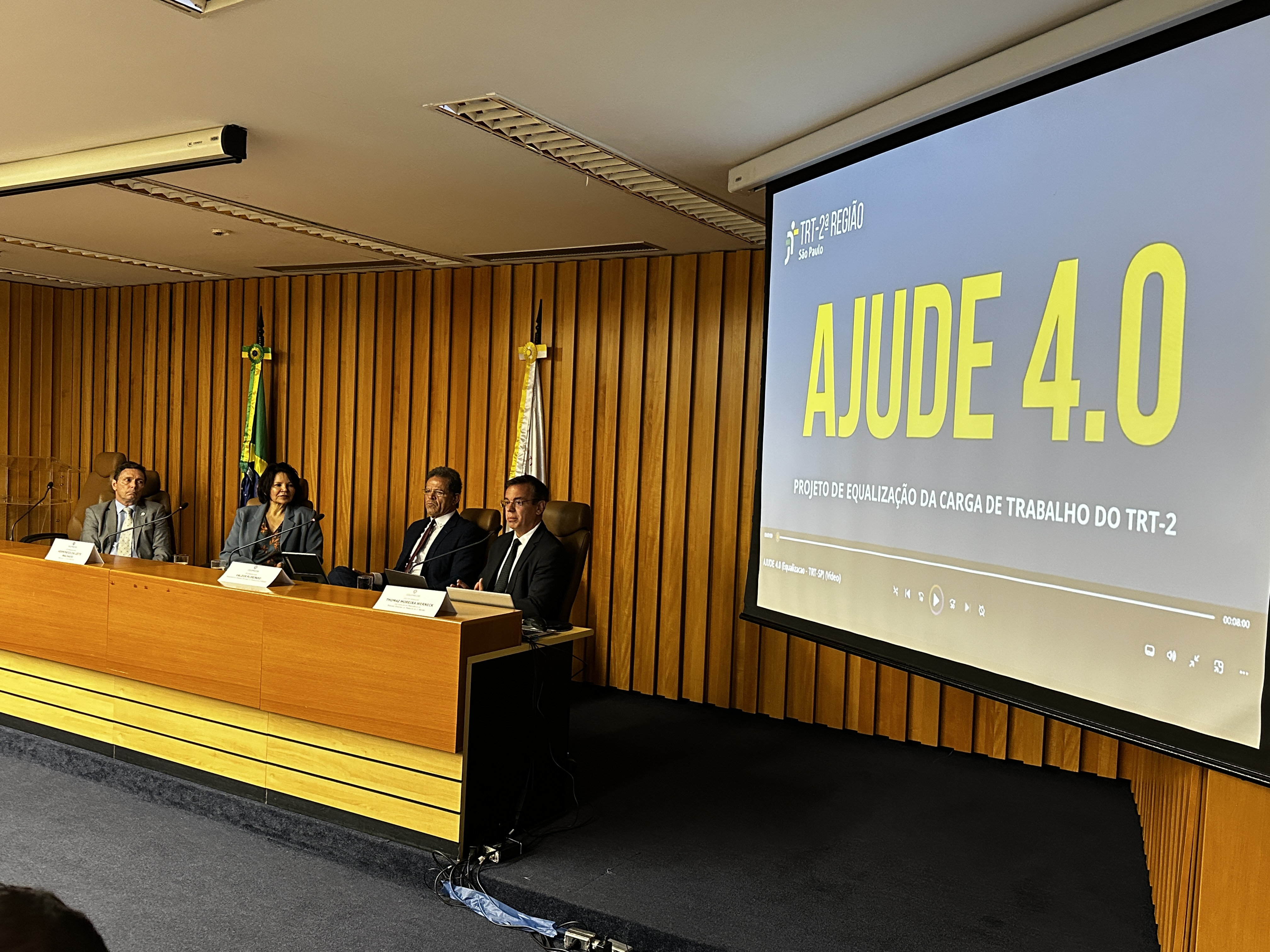A foto mostra pessoas numa mesa de autoridades ao lado de um telão.