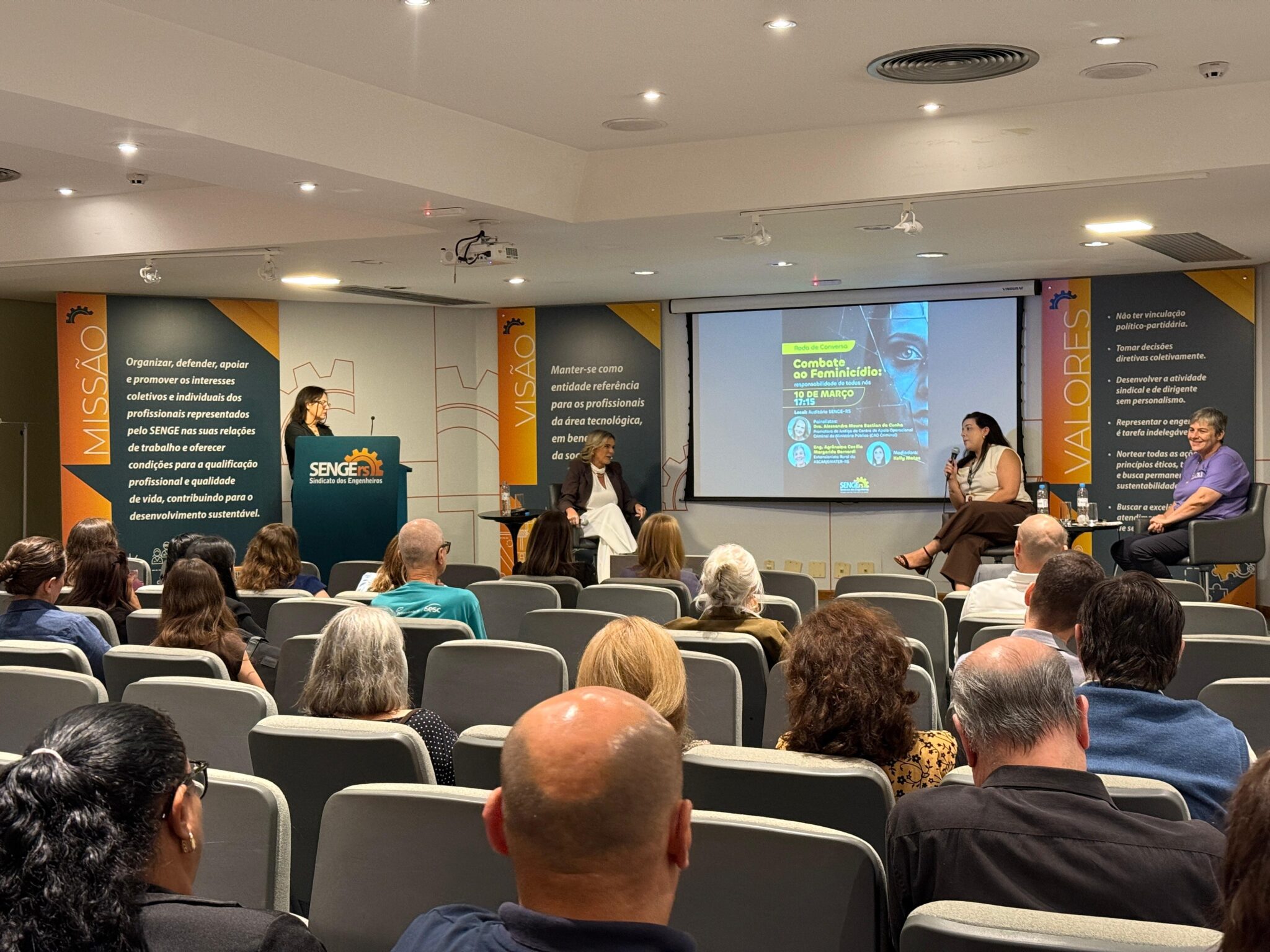 público em primeiro plano de costas, e ao fundo quatro mulheres aparecem debatendo no palco