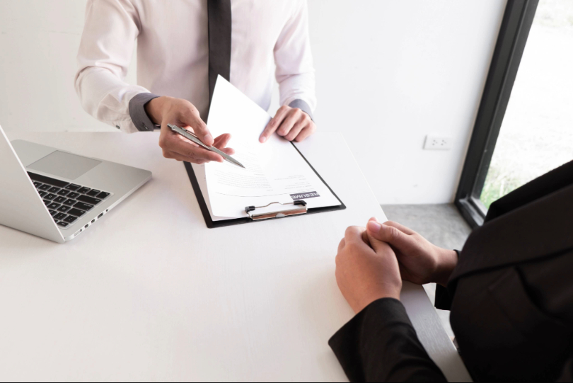 Entrevista de emprego: dois homens conversam. Um deles avalia um currículo sobre a mesa de escriório, enquanto o outro aguarda, com as mãos cruzadas. Não há imagem dos rostos. Um deles veste camisa branca e o outro, terno preto. Há um notebook prateado sobre a mesa.