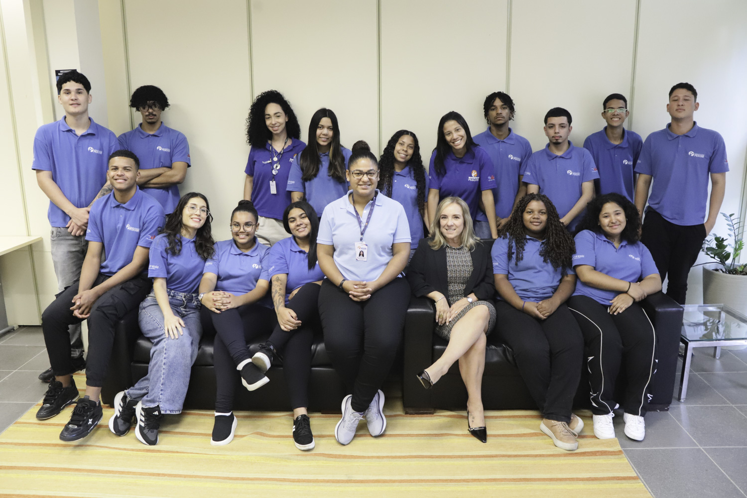 Turma de alunos posa para foto com a professora e a magistrada. Alguns estão sentados em sofás pretos e outros de pé. Os estudantes usam uniformes azuis do pescar.