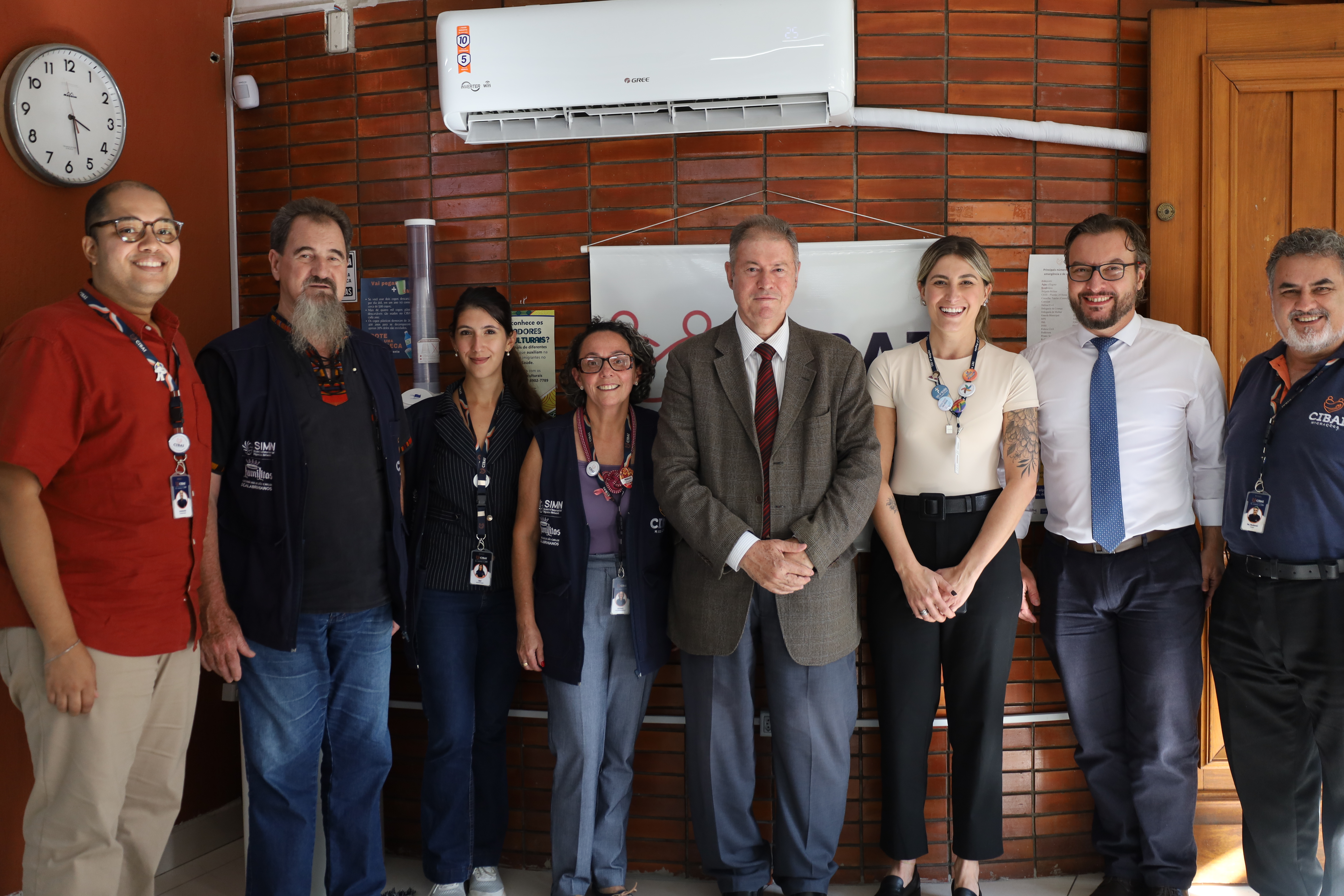 Equipe do CIBAI em um ambiente interno com parede de tijolos aparentes e um ar-condicionado branco no topo. À esquerda, um homem de camisa vermelha sorri, seguido por um senhor de barba longa e colete escuro. No centro, duas mulheres e dois homens em trajes formais (ternos e blazers) compõem o grupo principal, enquanto um homem de camisa polo azul fecha a fila à direita. 