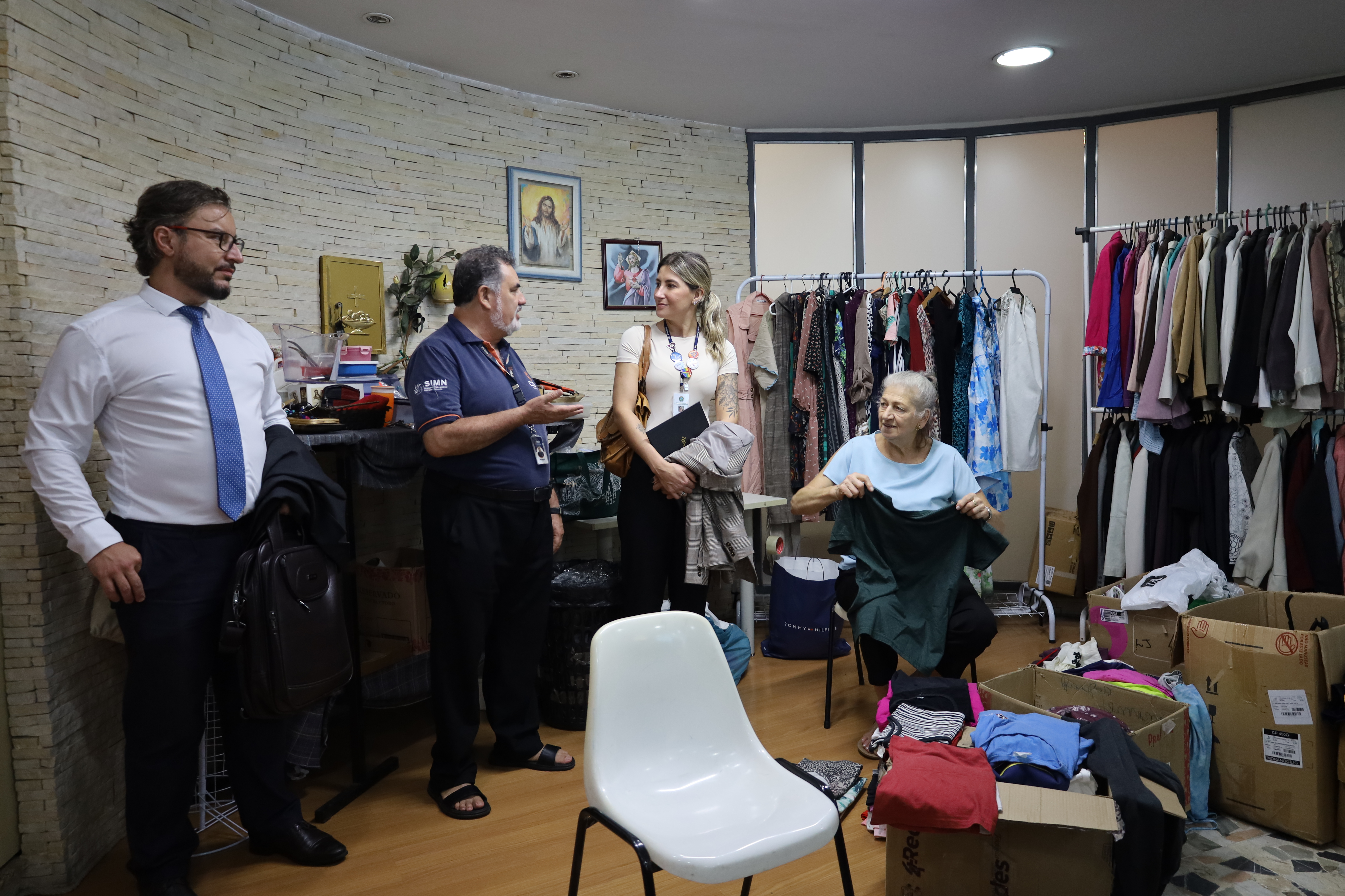 Em uma sala com paredes de textura de pedra, quatro pessoas interagem em um espaço destinado a doações. Uma mulher idosa está sentada ao centro, organizando uma peça de roupa verde sobre caixas de papelão repletas de vestimentas. À sua frente, os desembargadores de terno e uma servidora (anita) com blazer conversam com um funcionário de camisa polo azul que gesticula explicativamente. Ao fundo, araras repletas de roupas penduradas e quadros religiosos.