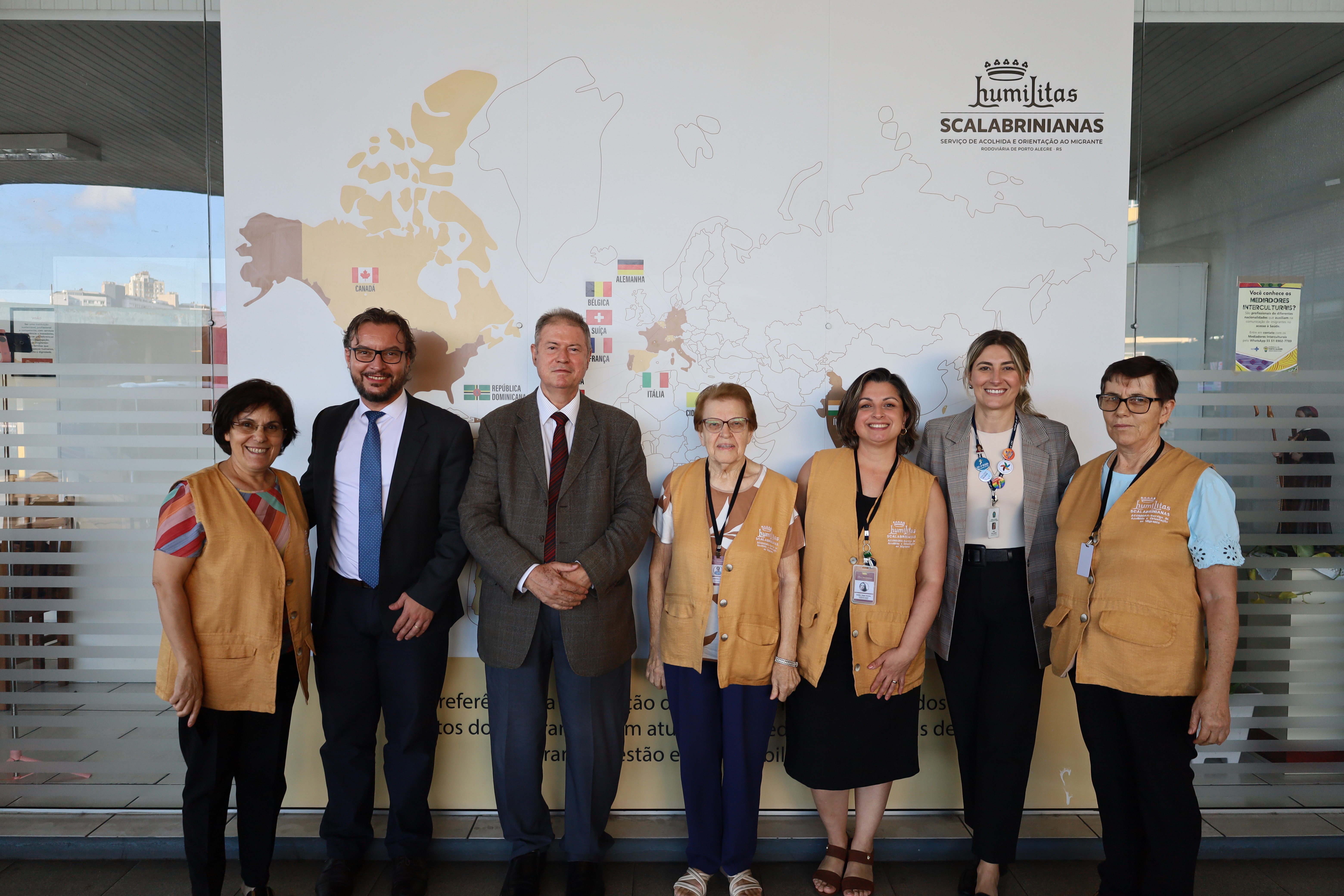 O grupo de sete pessoas posa diante de um grande painel branco que exibe um mapa-múndi e o logotipo "Humilitas Scalabrinianas". Quatro irmãs vestem coletes bege padronizados sobre suas roupas casuais, indicando serem voluntárias ou funcionárias da instituição. Entre elas, dois homens de terno e gravata e uma mulher (anita) em traje executivo cinza completam a composição. O local é bem iluminado por luz natural vinda de uma área envidraçada à esquerda, transmitindo um tom de visita oficial.