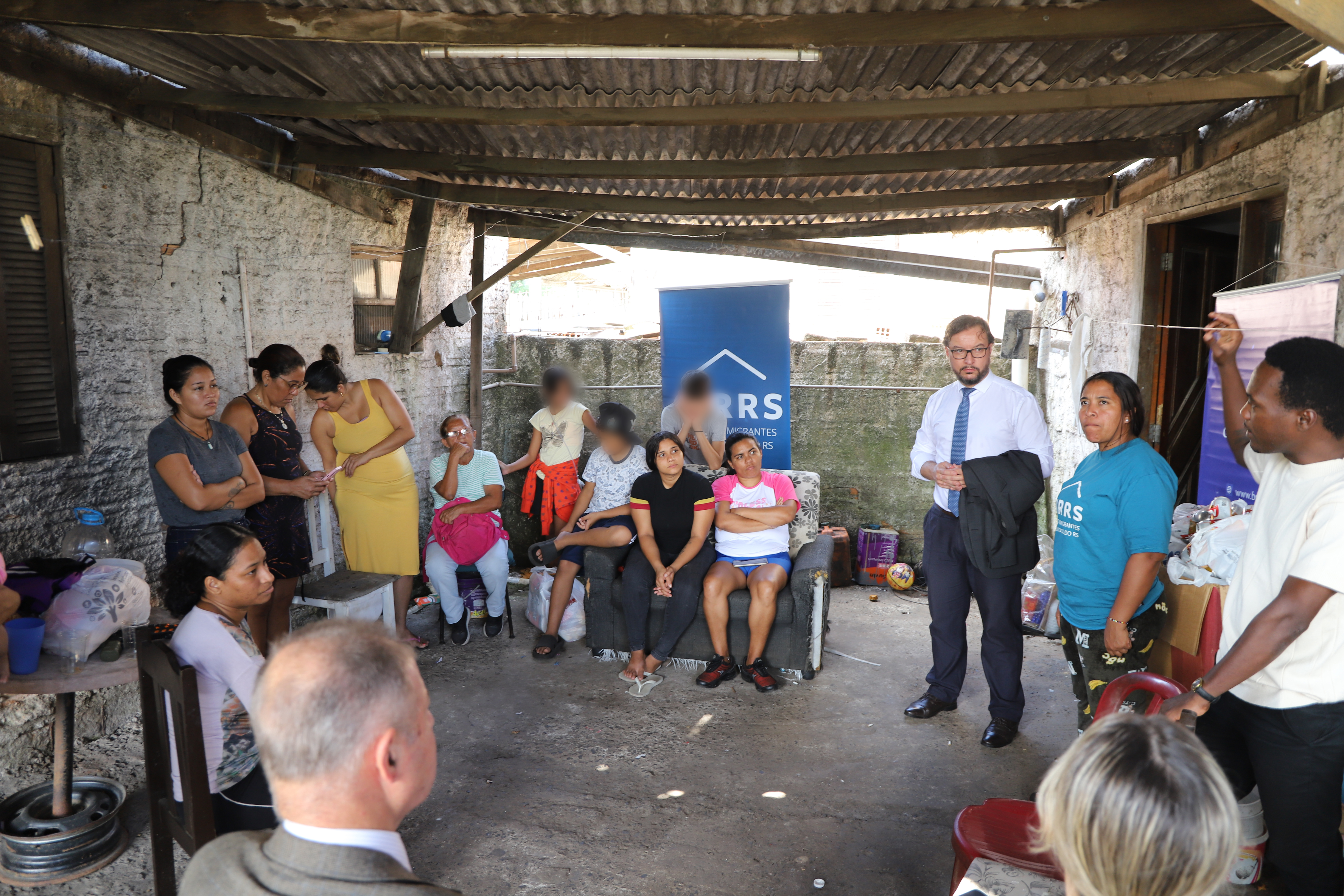 Encontro no Cirris. Um homem de camisa branca e gravata fala em pé, gesticulando para o grupo. Diversas mulheres e jovens ouvem atentamente sentados em um sofá antigo e cadeiras improvisadas. Ao fundo, nota-se um banner azul com a sigla "CIRRS".