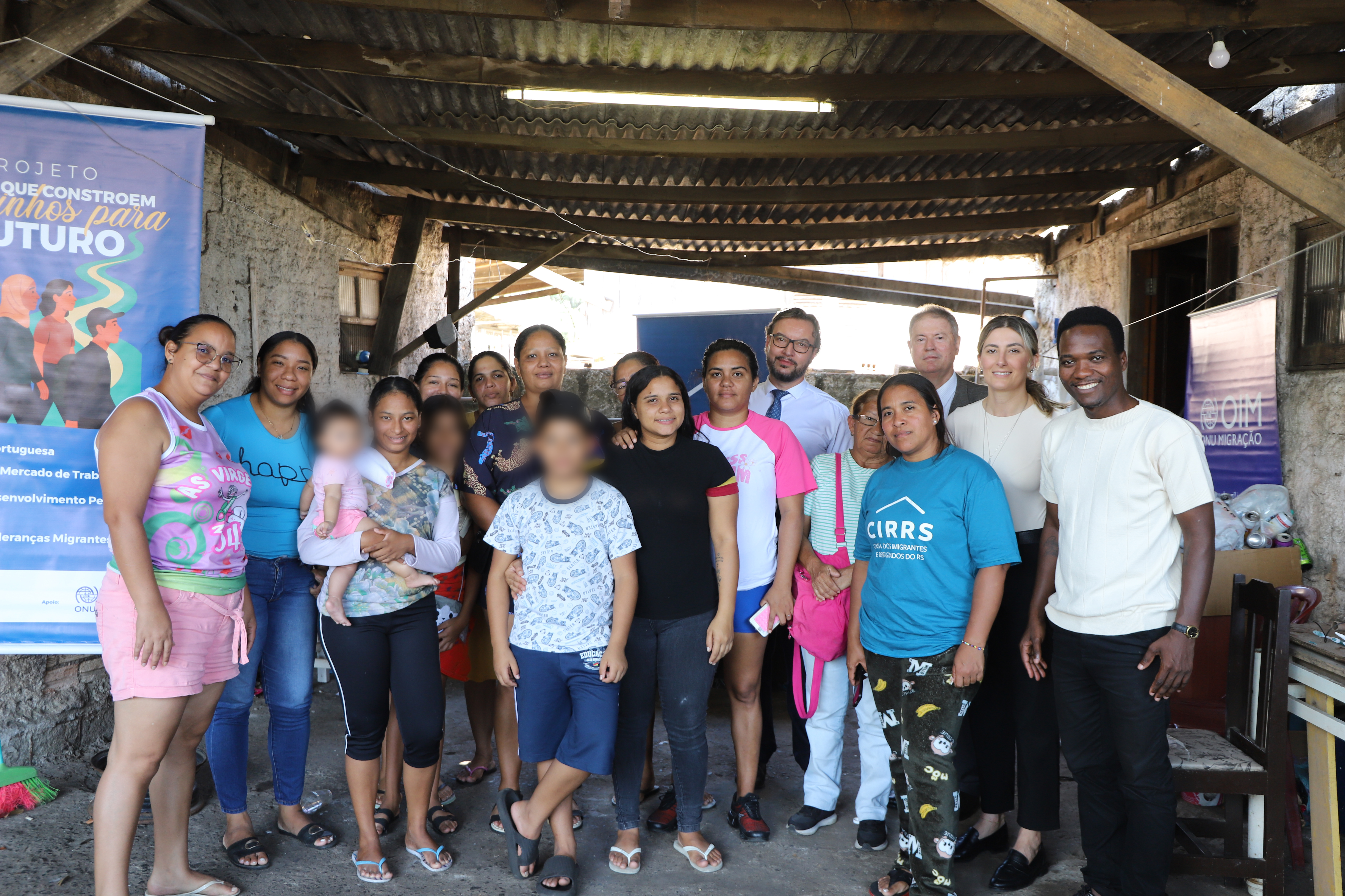 Um grupo numeroso posa para uma foto coletiva sob uma cobertura rústica de telhas onduladas e vigas de madeira. À esquerda, um banner colorido destaca o "Projeto Caminhos para o Futuro". No centro e à direita, os mesmos representantes formais das fotos anteriores aparecem integrados à comunidade atendida. O ambiente possui paredes de chapisco e chão batido, evidenciando uma ação realizada diretamente em território comunitário.