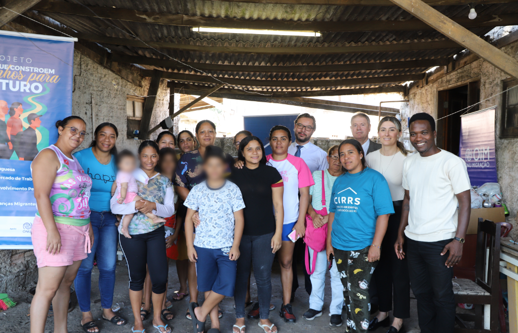 Um grupo numeroso posa para uma foto coletiva sob uma cobertura rústica de telhas onduladas e vigas de madeira. À esquerda, um banner colorido destaca o "Projeto Caminhos para o Futuro". No centro e à direita, os mesmos representantes formais das fotos anteriores aparecem integrados à comunidade atendida. O ambiente possui paredes de chapisco e chão batido, evidenciando uma ação realizada diretamente em território comunitário.