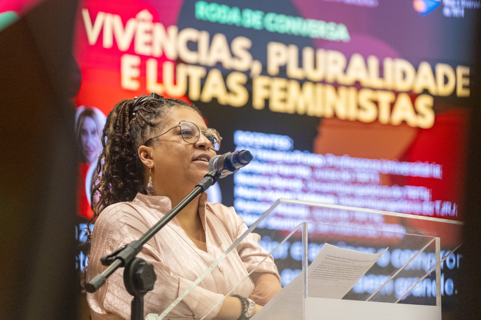 Vânia é negra e tem cabelos cacheados em tranças. Usa blusa rosa e fala ao microfone com o telão da atividade ao fundo