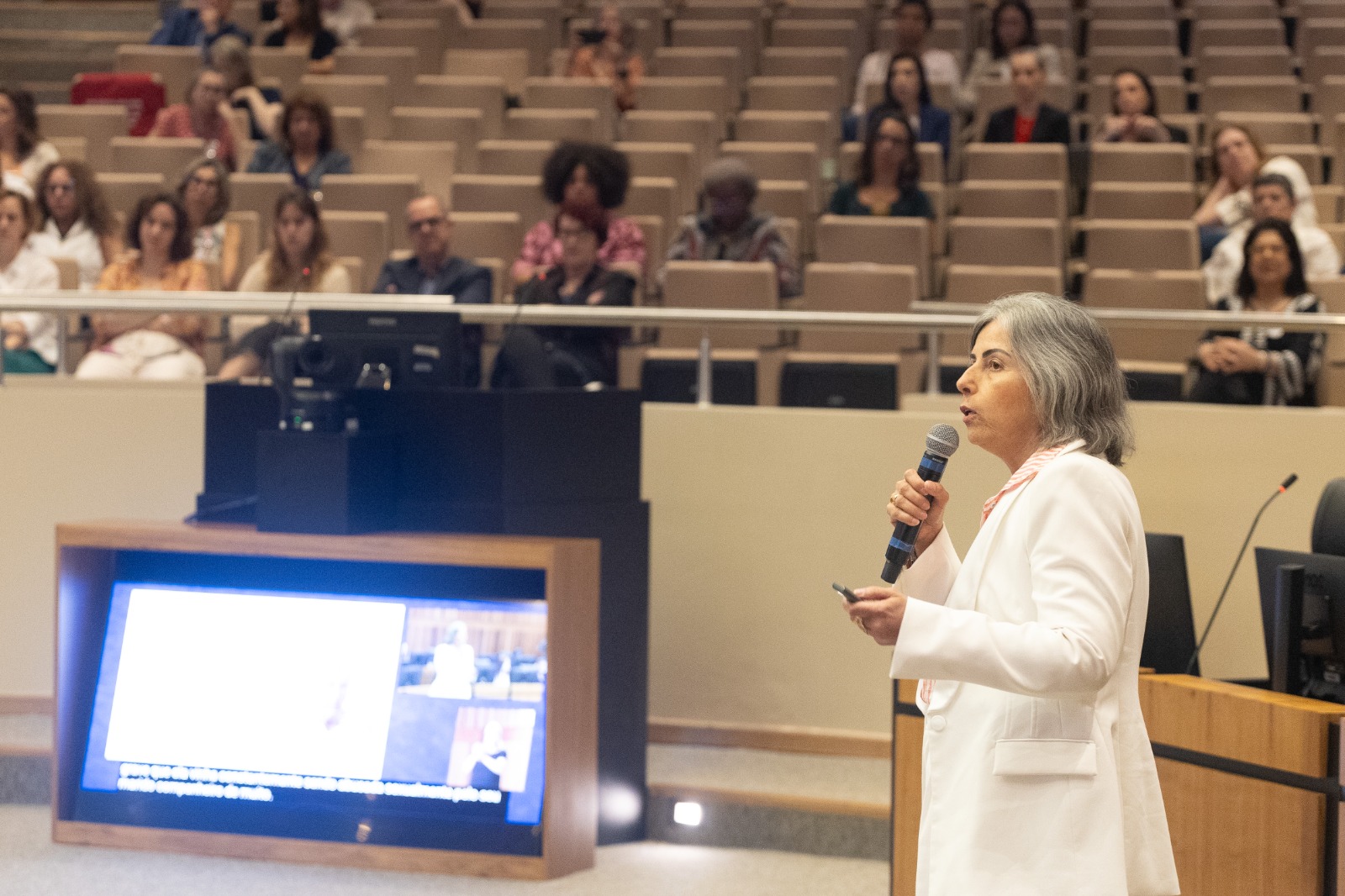 Maria é branca e grisalha, veste casaco branco. fala de pé no canto direito. ao fundo aparece o público. 
