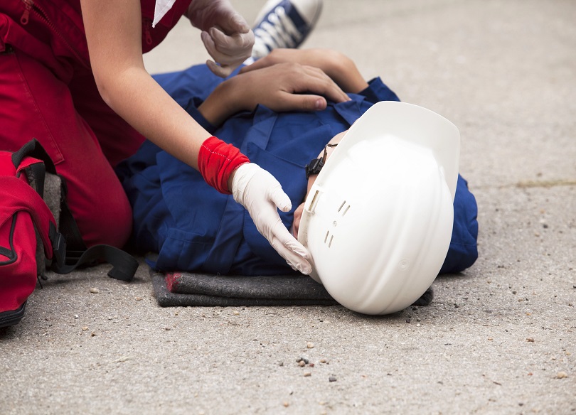 A foto ilustrativa mostra uma situação de atendimento de emergência. Um homem está deitado no chão, aparentemente inconsciente ou ferido, usando uniforme azul e um capacete branco de segurança, sugerindo que pode ser um trabalhador.