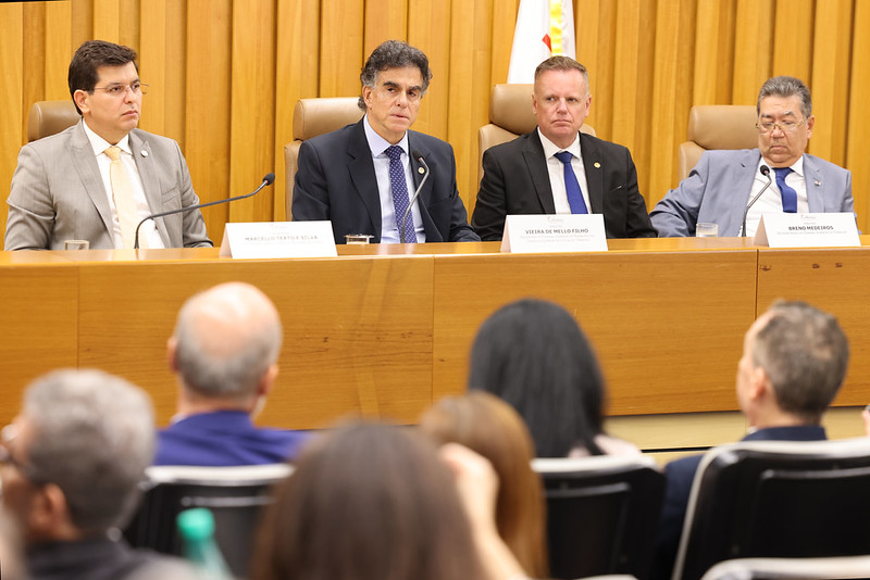 quatro pessoas falam na mesa oficial, com o público de costas. Vieira de Mello fala ao microfone. 