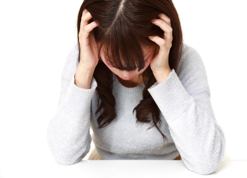 A foto ilustrativa mostra uma mulher sentada, com a cabeça inclinada para baixo e as mãos apoiadas nas laterais da cabeça, como se estivesse segurando ou pressionando o couro cabeludo. O rosto dela não está totalmente visível, pois o cabelo cobre parte da face.