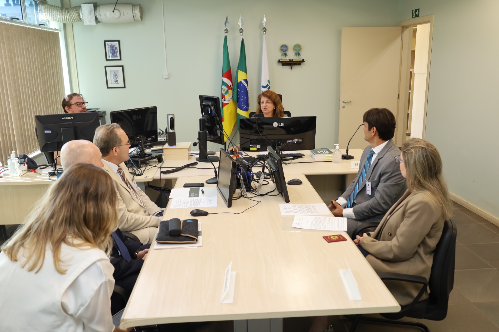Imagem mostra, de forma centralizada, juíza, assistente de secretaria, partes e procuradores, sentados, conversando, em sala de audiências. A juíza está ao centro da fotografia, do lado esquerdo está a parte reclamante, e do lado direito está a reclamada