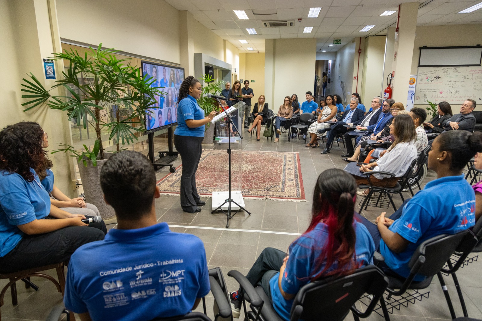 aluna de pé ao  centro fala para a plateia que senta-se em semicírculo