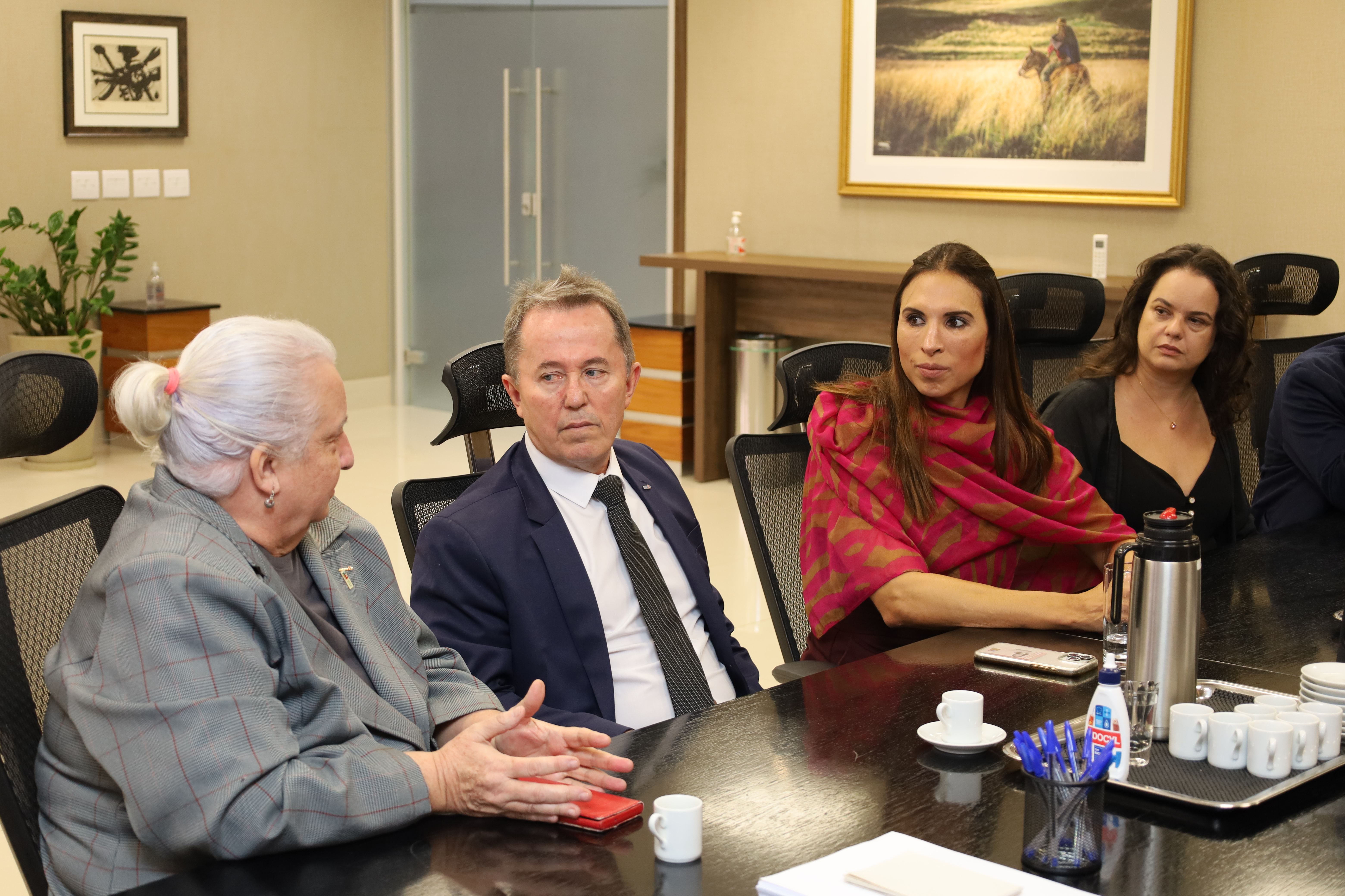 A fotografia mostra os participantes da reunião, sentados ao redor da mesa oval, conversando.