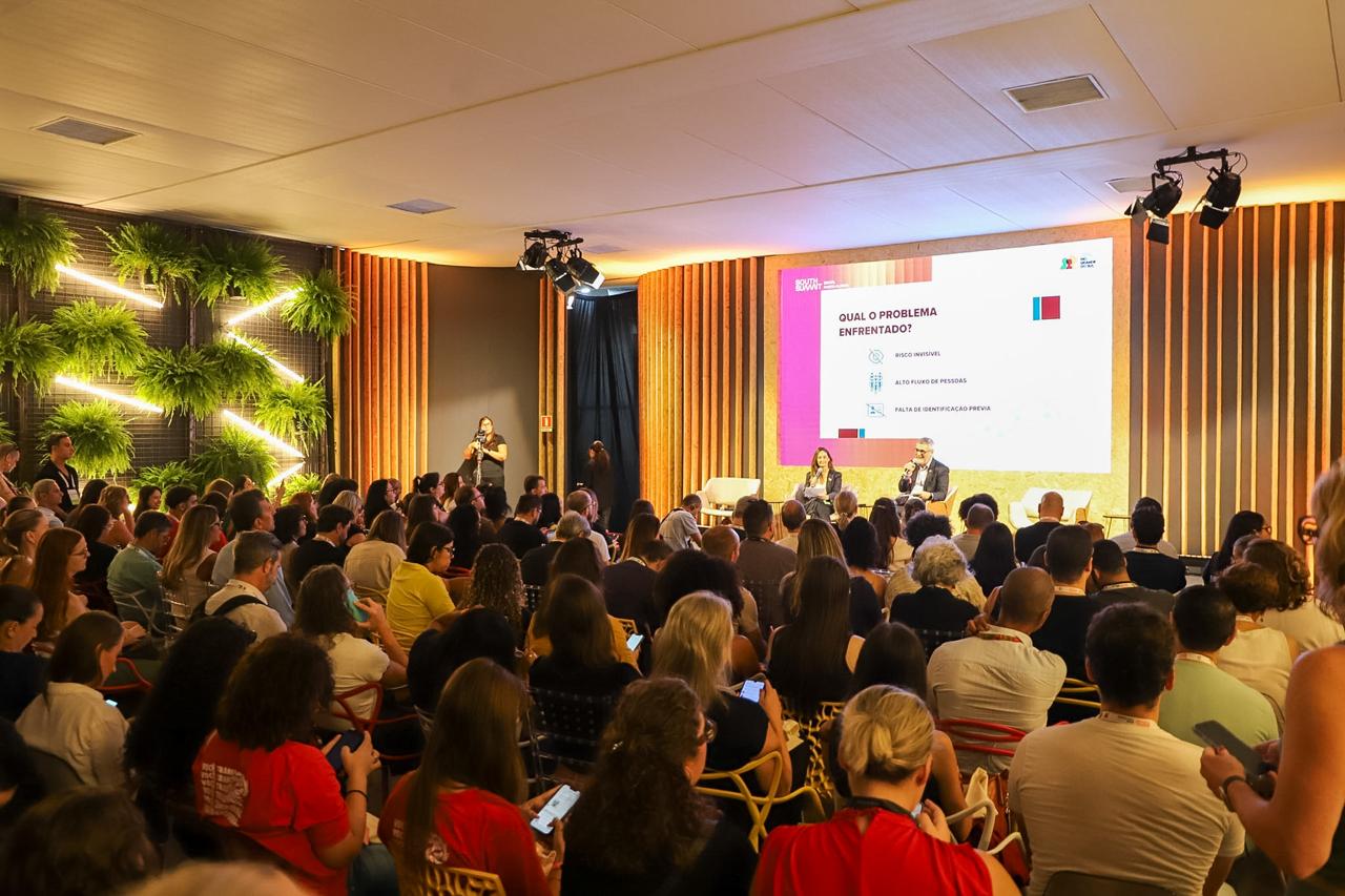 Foto em ângulo aberto, mostrando auditório lotado e o palco ao fundo. No palco, a juíza Luciana e o servidor Peixoto
