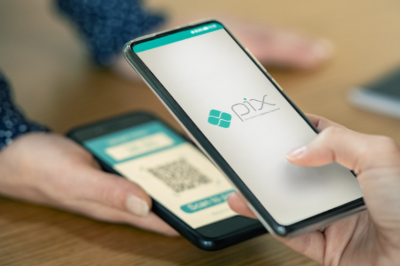 Duas mãos seguram smartphones sobre uma superfície de madeira, com um dos aparelhos exibindo o logotipo do Pix enquanto se posiciona para escanear um código QR no outro celular.