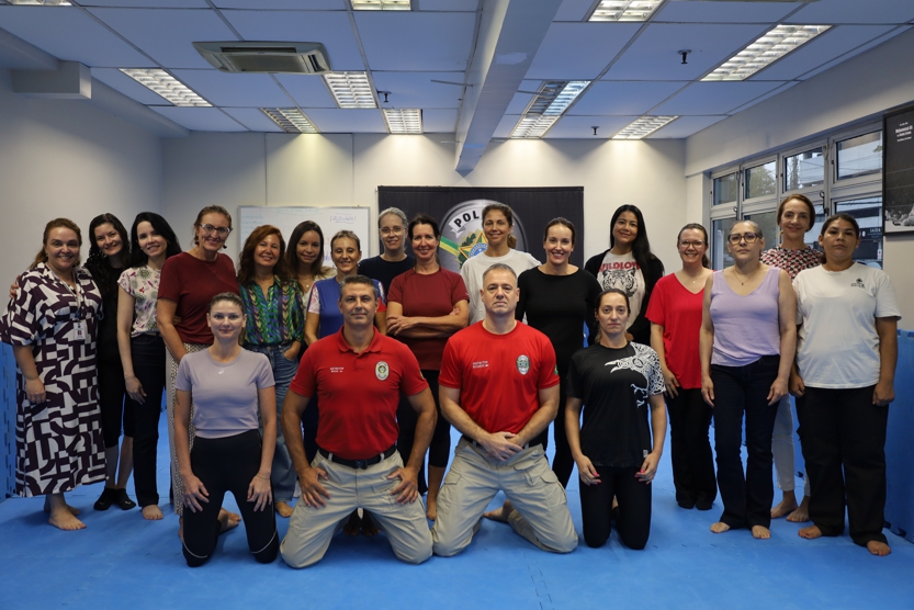 Grupo de participantes reunido em uma sala de treinamento com piso acolchoado azul. Na primeira fila, três pessoas estão ajoelhadas, incluindo dois instrutores com camisetas vermelhas da Polícia Judicial e uma participante ao lado. Atrás, uma fileira de mulheres em pé forma o restante do grupo, com roupas variadas, entre trajes casuais e esportivos.