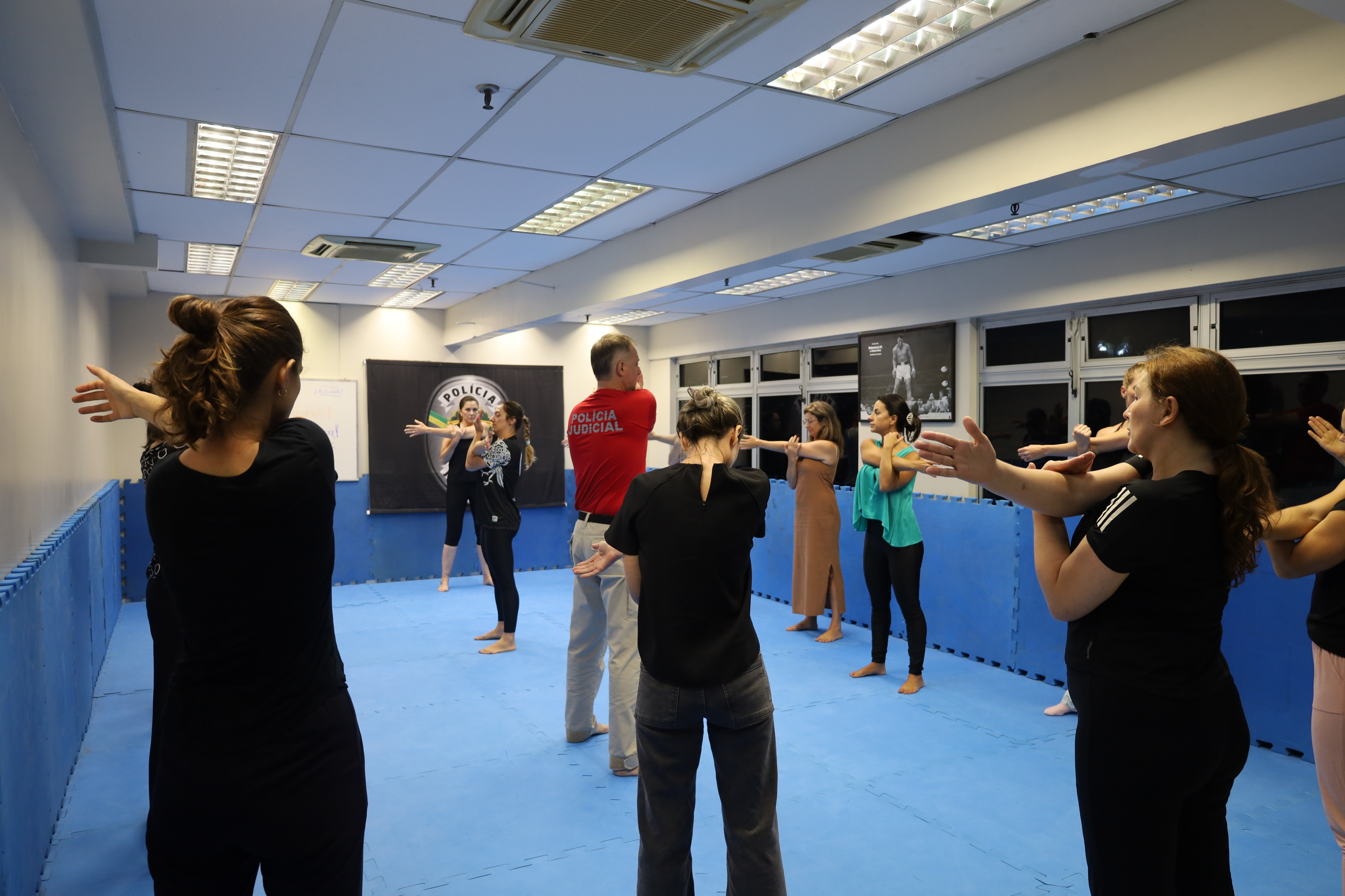 Um instrutor, ao centro, orienta um grupo de mulheres dispostas em círculo. Todas executam alongamentos com os braços estendidos. Ao fundo, há um banner da Polícia Judicial. As participantes usam roupas confortáveis, adequadas para atividade física, e estão descalças, focadas na prática.