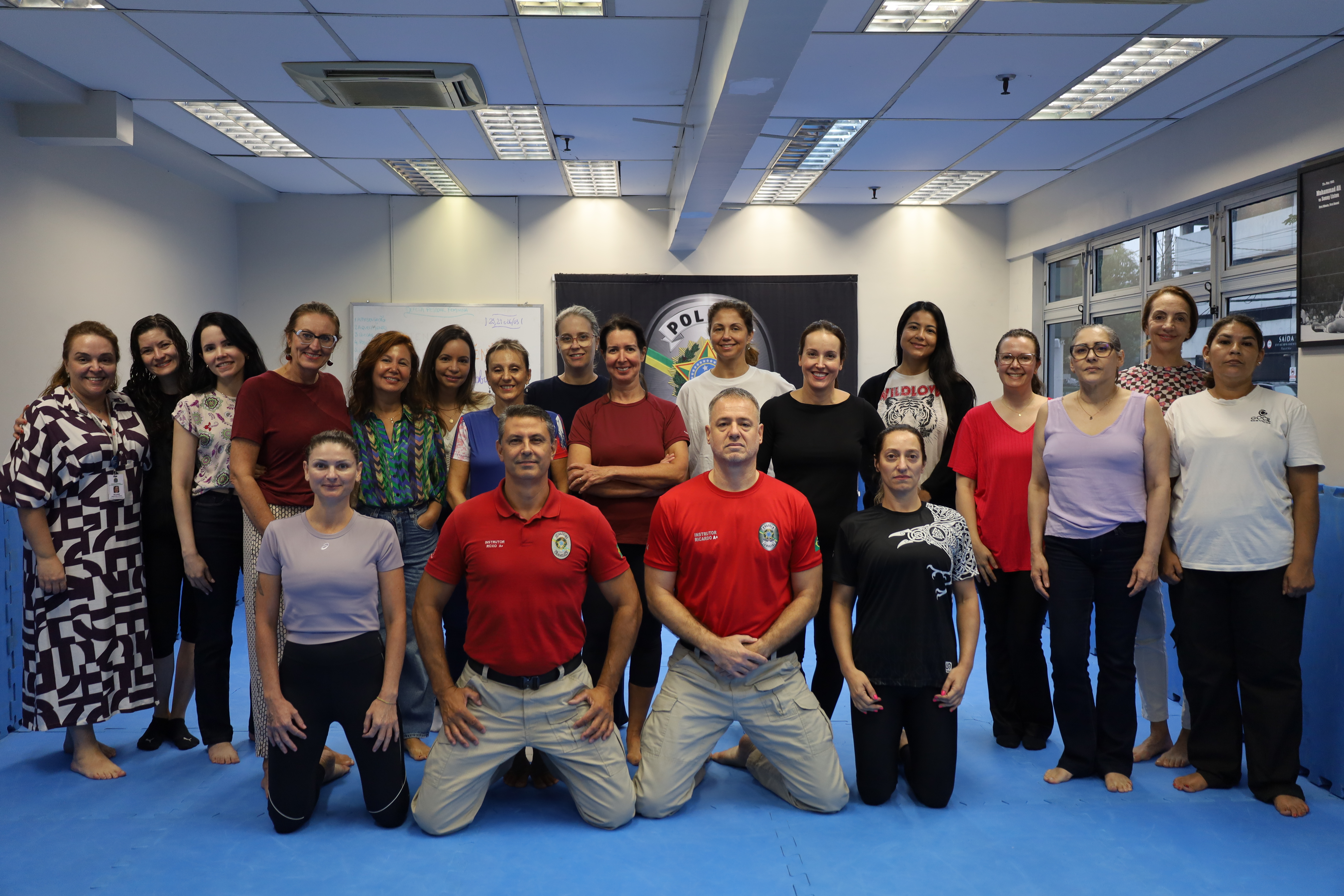 Grupo de participantes reunido em uma sala de treinamento com piso acolchoado azul. Na primeira fila, três pessoas estão ajoelhadas, incluindo dois instrutores com camisetas vermelhas da Polícia Judicial e uma participante ao lado. Atrás, uma fileira de mulheres em pé forma o restante do grupo, com roupas variadas, entre trajes casuais e esportivos. Ao fundo, há um banner da Polícia Judicial e um quadro branco.